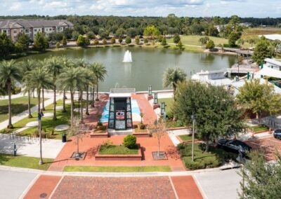 Aerial view of a landscaped park with palm trees, a red brick path, a central fountain in a pond, and surrounding residential and commercial buildings under a partly cloudy sky.