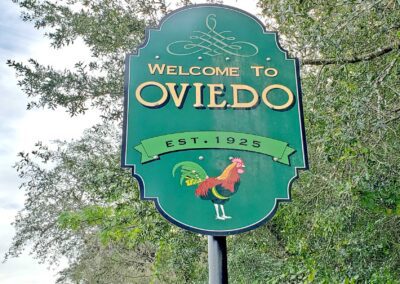 Green sign reading "Welcome to Oviedo, Est. 1925" with an illustration of a rooster. Trees are visible in the background.