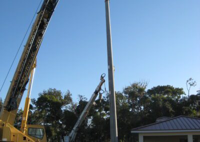 Cranes installing a tall light pole on a grassy field near a small building under a clear blue sky.