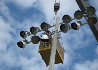 A worker in a yellow lift repairs a tall stadium light tower on a cloudy day.