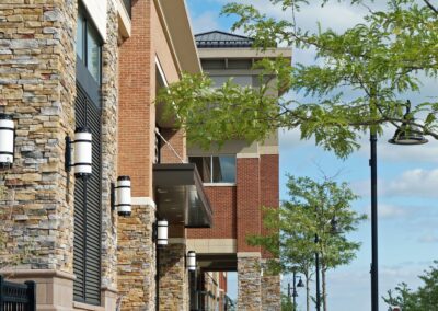 Sidewalk view with modern brick buildings, stone accents, black benches, and trees lining the street under a partly cloudy sky.